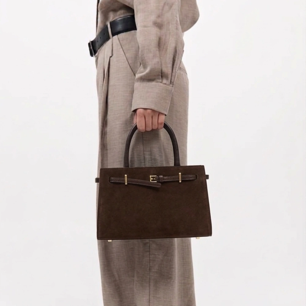 Woman holding Espresso Suede Mini Book Tote in dark chocolate brown suede with gold hardware and Nappa calf leather base, styled with tailored neutral trousers and black belt
